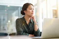 A young woman sits thoughtfully at a sunlit office desk, resting her chin on her hand while looking away from her open laptop. AI generated.