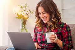 A smiling woman with wavy brown hair sitting comfortably on a couch in a brightly lit room, wearing a red plaid shirt. She is holding a white mug and looking happily at a laptop resting on her lap, with a vase of white flowers glowing in the sunlight behind her. AI generated.