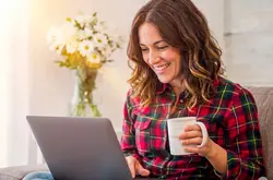 A smiling woman with wavy brown hair sitting comfortably on a couch in a brightly lit room, wearing a red plaid shirt. She is holding a white mug and looking happily at a laptop resting on her lap, with a vase of white flowers glowing in the sunlight behind her. AI generated.
