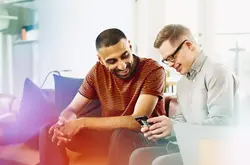 Two people sitting on a couch smiling while looking at a phone.
