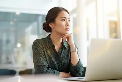 A young woman sits thoughtfully at a sunlit office desk, resting her chin on her hand while looking away from her open laptop. AI generated.