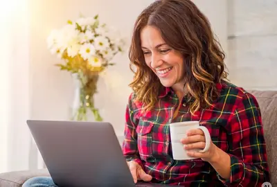 A smiling woman with wavy brown hair sitting comfortably on a couch in a brightly lit room, wearing a red plaid shirt. She is holding a white mug and looking happily at a laptop resting on her lap, with a vase of white flowers glowing in the sunlight behind her. AI generated.