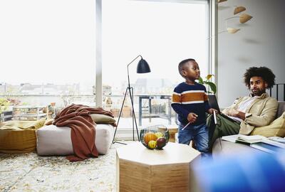 Man sitting on couch, working on laptop. Kid holding a notebook and pen.