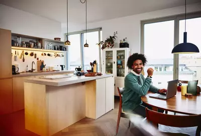 Smiling male sitting at his dining table with his laptop. Kitchen in the background.
