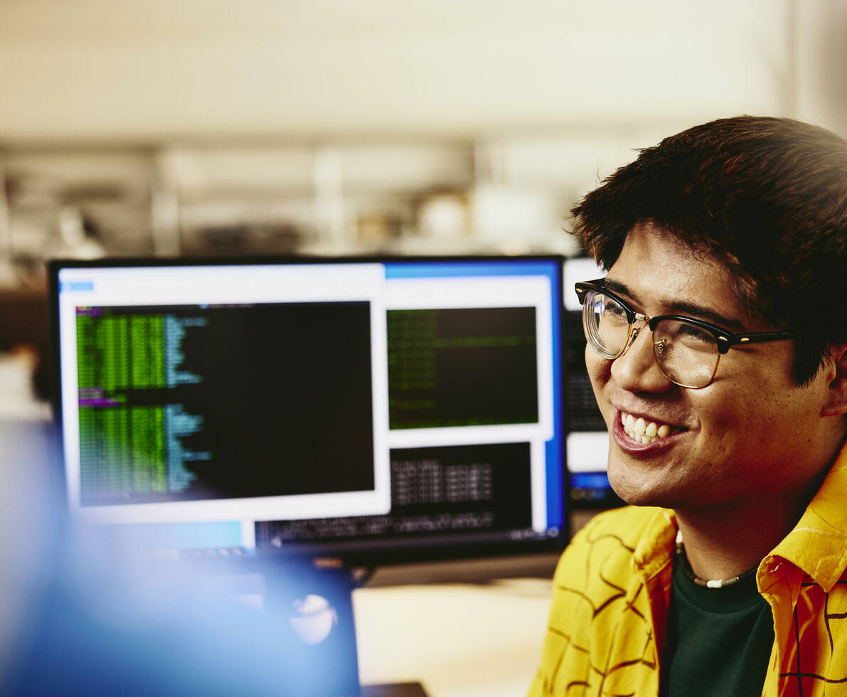 Man smiling while sitting behind his desk, computer screens displaying programming code.