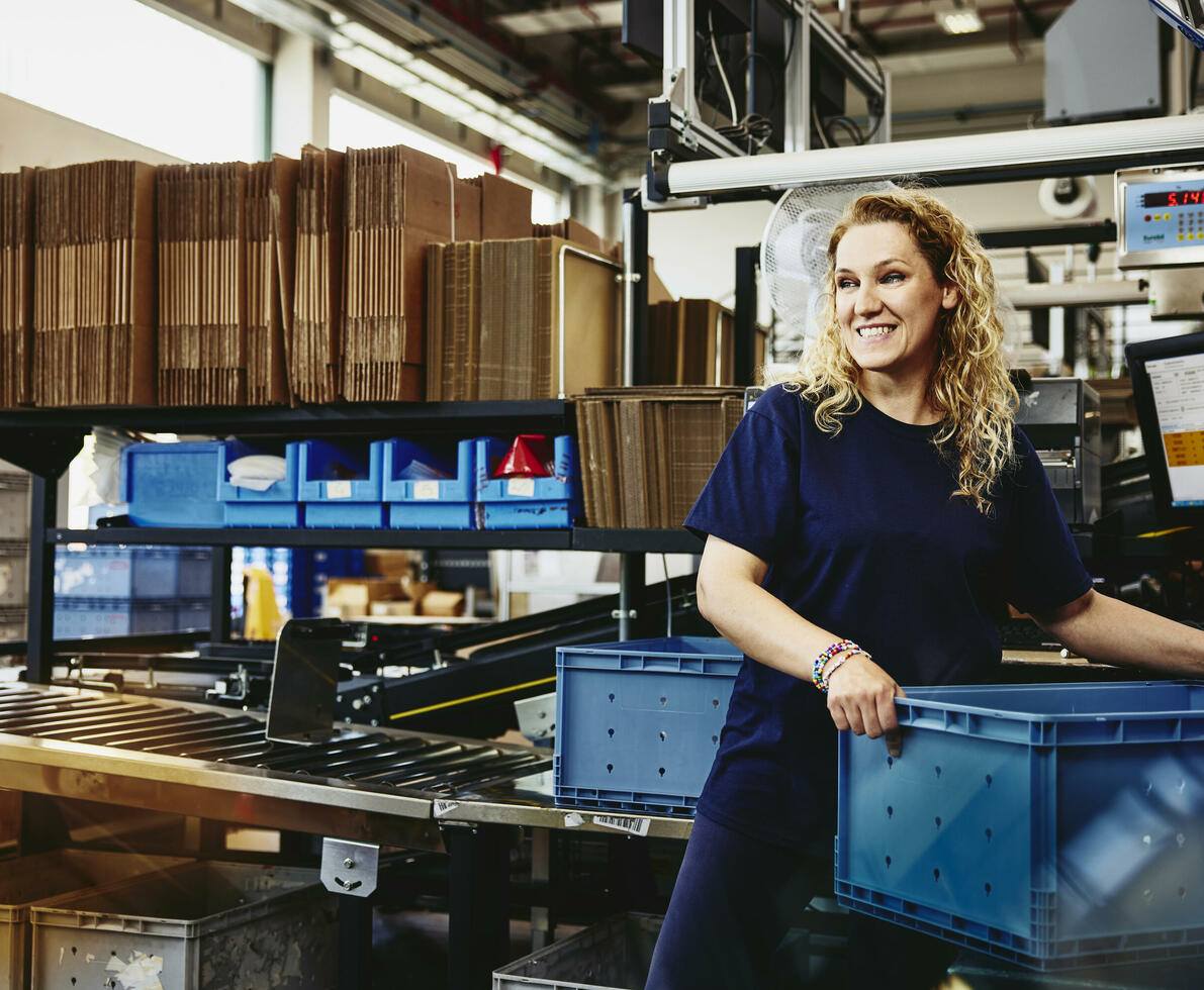 Smiling woman standing next to conveyer belt holding a crate