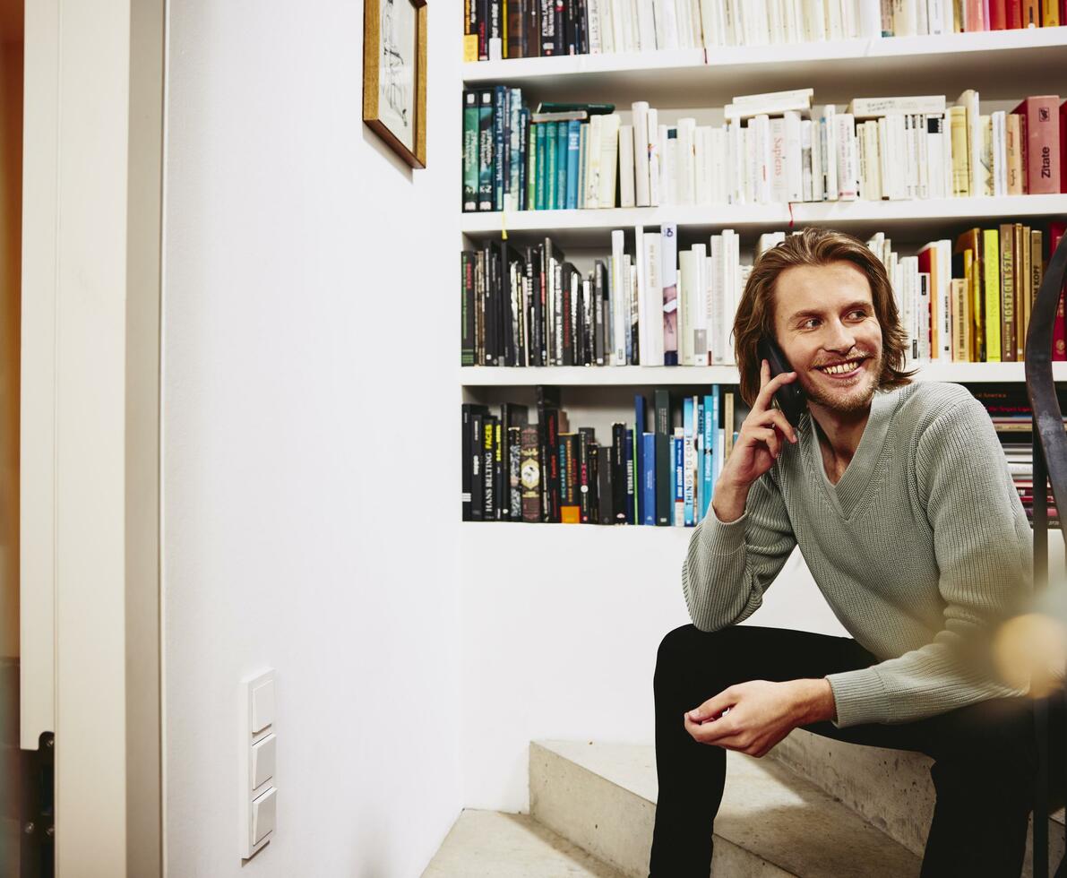 Smiling man sitting on stairs, having a phonecall. bookshelf in background