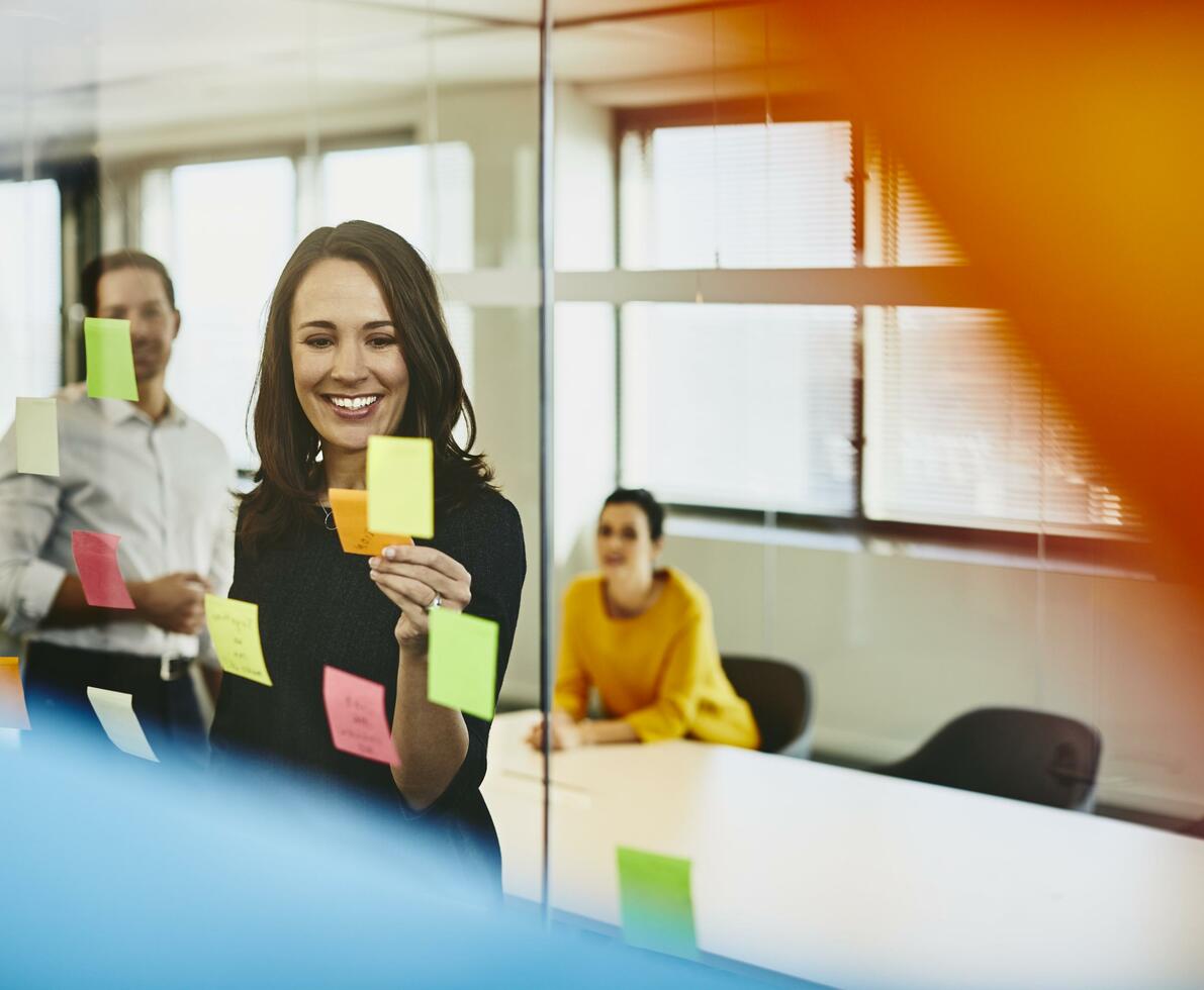 Two business women and a man in an office putting sticky notes on a window. Primary colors: yellow and blue.