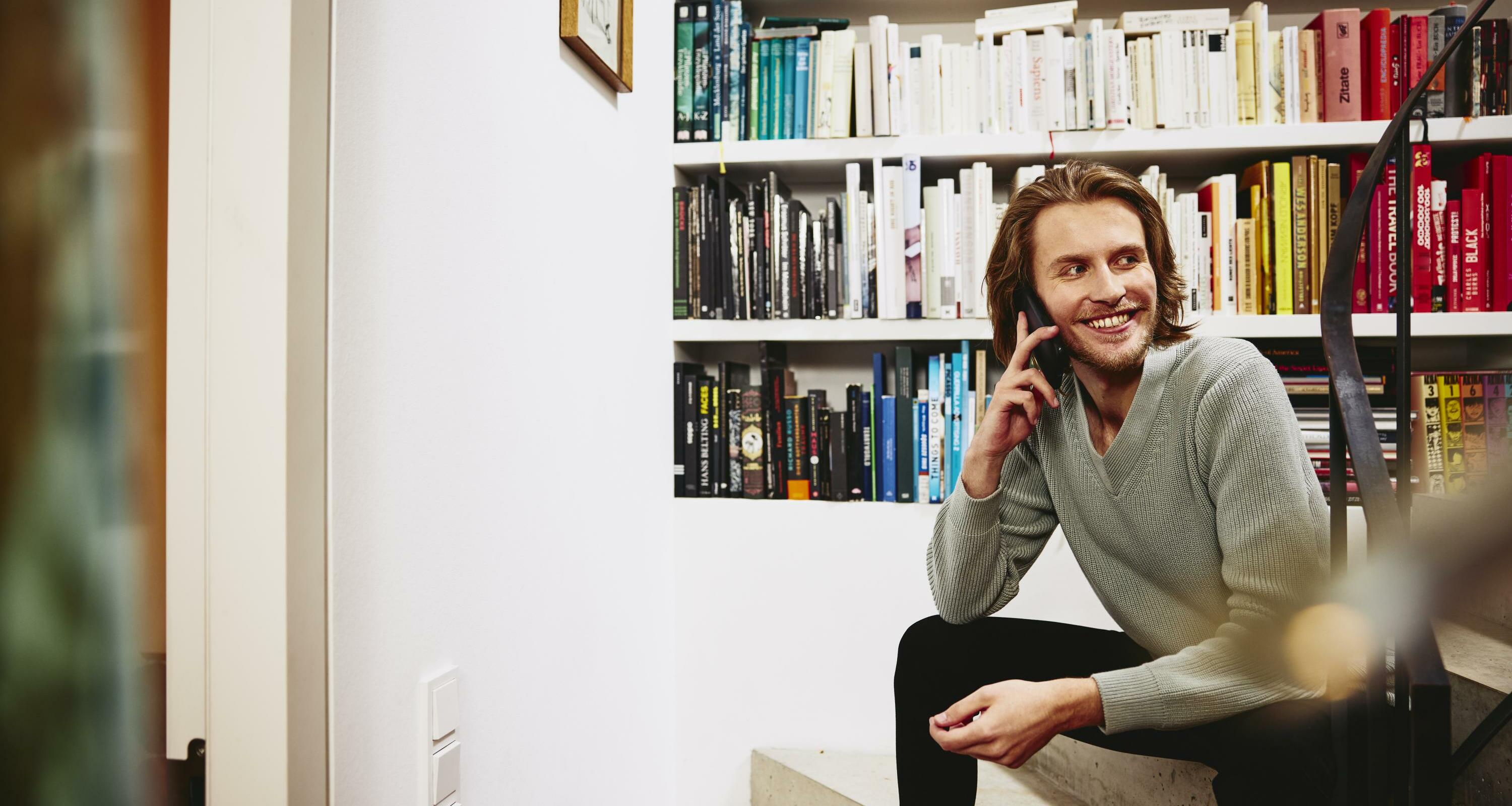Smiling man sitting on stairs, having a phonecall. bookshelf in background