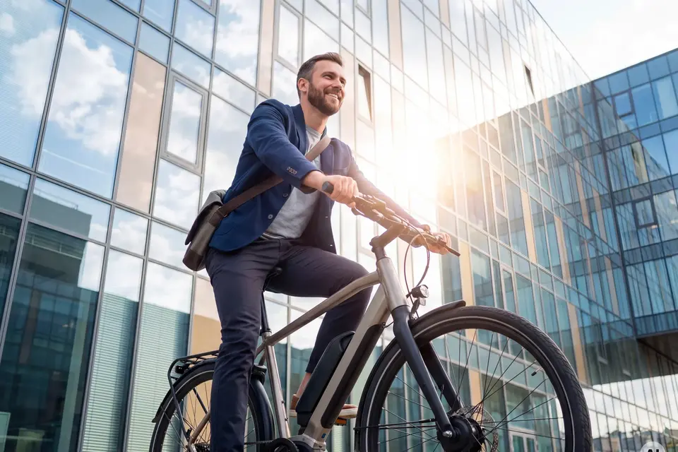A smiling man wearing a blue blazer and a messenger bag rides an electric bicycle past a modern glass office building on a sunny day. AI generated.